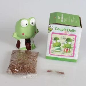 A small green frog-shaped planter kit is displayed on a white background. The frog figurine, wearing a white shirt with red suspenders, sits next to a green-and-white product box labeled “Couple Dolls.” In front of the figurine is a sealed packet of growing medium or soil and a small packet of seeds. The box shows illustrations of two frog planters with grass growing from their heads.