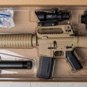 Overhead view of a desert tan M-16 gel blaster rifle resting in a clear plastic molded tray inside a cardboard box. The kit includes a black magazine, an ACOG-style scope, a red dot sight, a black suppressor, and a pair of blue safety glasses protected by bubble wrap.
