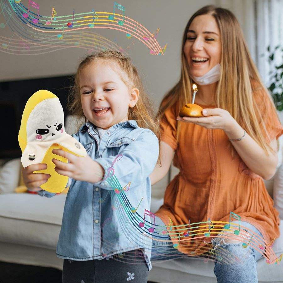 Fruit body sensing music piano plush Banana strawbery carrot fun toy for kids buy online Beirut Lebanon hand eye coordination A young child laughing while holding and interacting with a plush banana music piano body sensory toy that features a cute, expressive face, while a woman sits behind them holding a small cake with a candle, and graphic overlays of colorful musical notes swirl around them to represent the interactive sound features of the toy.