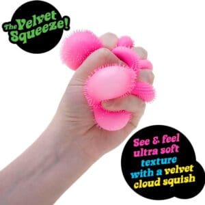 A hand squeezing a bright pink, fuzzy sensory toy against a plain white background. Black graphic bubbles feature the text "The Velvet Squeeze!" and "See & feel ultra soft texture with a velvet cloud squish."