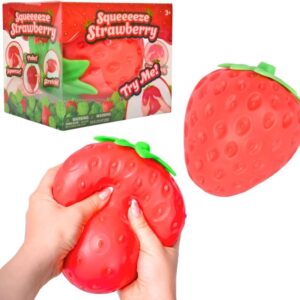 Bright red soft strawberry squishy stress toy being squeezed by hands next to its packaging box, available for online buy in Beirut, Lebanon. Arabic: لعبة فراولة مطاطية ناعمة زاهية اللون يتم ضغطها باليدين مع علبة التغليف الخاصة بها، متوفرة للشراء أونلاين في بيروت، لبنان
